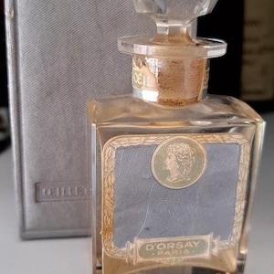 Peut inclure: Un flacon de parfum vintage avec un bouchon en cristal facetté. La bouteille porte une étiquette avec un cadre doré et un portrait de femme. Le texte "D'ORSAY PARIS" est visible. Un étui gris est en arrière-plan.