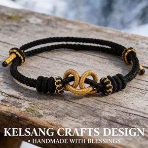 Può includere: Bracciale nero e oro con un design a doppio nodo infinito. Il bracciale presenta un cordino nero con dettagli dorati e un nodo centrale dorato. Il testo "KELSANG CRAFTS DESIGN - HANDMADE WITH BLESSINGS" è in basso.