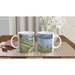 Pode incluir: Duas canecas de cerâmica branca com uma ilustração de paisagem contínua. O design mostra uma colina relvada com um caminho de terra sinuoso, pinheiros dispersos e um lago azul calmo sob montanhas distantes com flores silvestres.