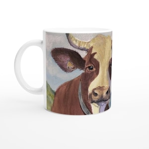 May include: White ceramic mug with a watercolour painting of a brown and white cow's face. The cow has a tag in its ear with the number 8368. The background shows a light blue sky and green hills.