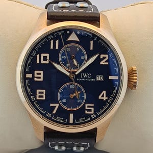 May include: A rose gold wristwatch with a dark blue face, featuring white hands and numerals. The watch has a black leather strap and the brand name "IWC SCHAFFHAUSEN" is visible. The watch is displayed on a cream-coloured surface.
