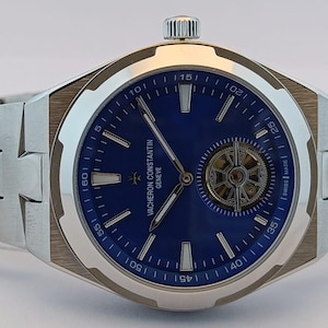 May include: A stainless steel wristwatch with a blue dial and a visible escapement. The watch features silver hands and markers, and a silver band. The watch face has the words "Vacheron Constantin Geneve" printed on it.