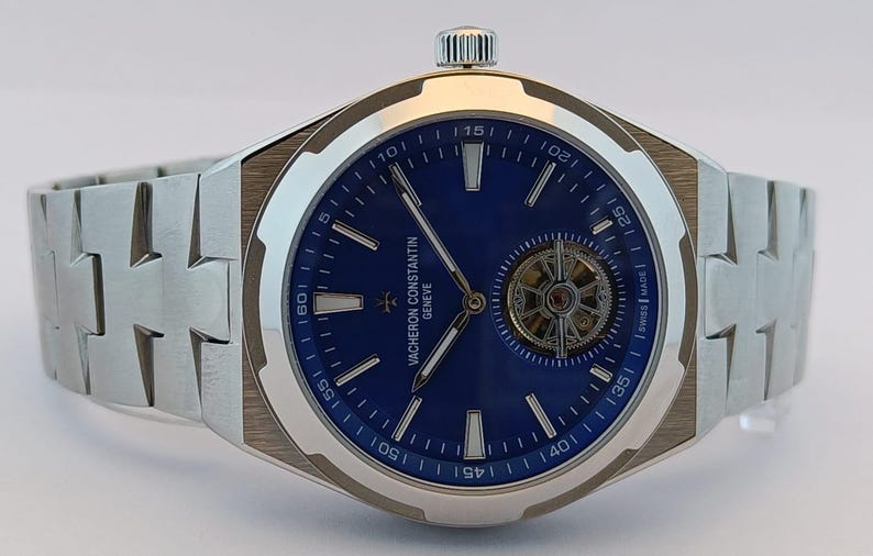 May include: A stainless steel wristwatch with a blue dial and a visible escapement. The watch features silver hands and markers, and a silver band. The watch face has the words "Vacheron Constantin Geneve" printed on it.