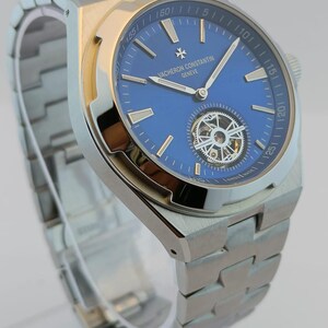 May include: A stainless steel wristwatch with a blue dial. The watch features a silver band and a visible mechanical movement. The dial includes the text "VACHERON CONSTANTIN GENEVE".