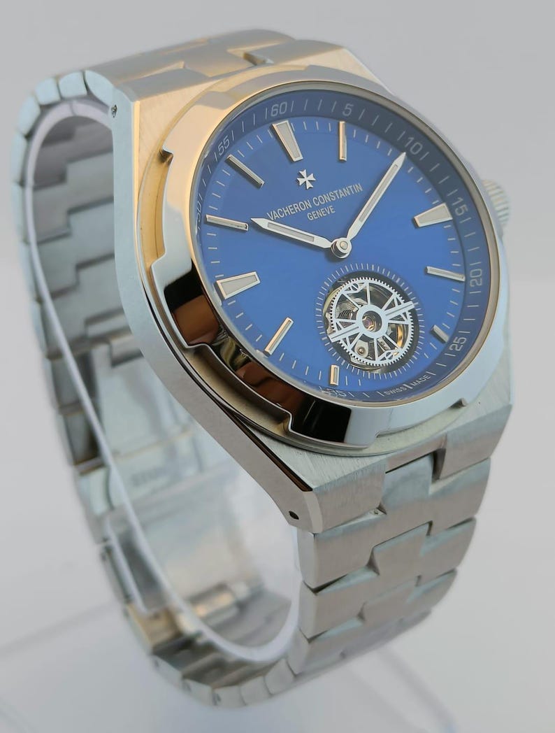 May include: A stainless steel wristwatch with a blue dial. The watch features a silver band and a visible mechanical movement. The dial includes the text "VACHERON CONSTANTIN GENEVE".
