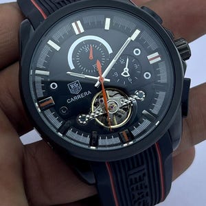 Op de afbeelding: Zwarte Carrera-horloge met een zwarte wijzerplaat, witte accenten en oranje wijzers. Het horloge heeft een zwarte rubberen band met rode strepen. De wijzerplaat toont meerdere sub-wijzerplaten en het Carrera-logo.