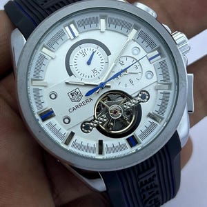 Op de afbeelding: Een zilverkleurig Carrera horloge met een witte wijzerplaat, blauwe accenten en een zwarte rubberen band. Het horloge heeft meerdere sub-wijzerplaten en een zichtbaar mechanisch uurwerk. De band heeft het woord "EAGLE" in reliëf.