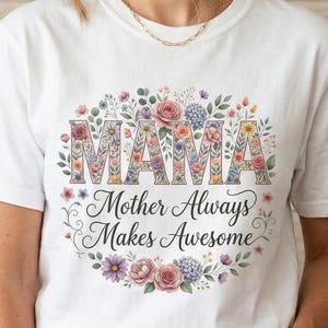 Puede incluir: Camiseta blanca con la palabra "MAMA" en letras florales, rodeada por una corona floral. El texto "Mother Always Makes Awesome" está debajo. La modelo lleva pantalones cortos vaqueros azules.