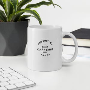 May include: A white ceramic coffee mug with a black graphic that reads "Powered by Caffeine and AI." The mug has a curved handle and is on a white surface. A plant in a black pot and a keyboard are in the background.