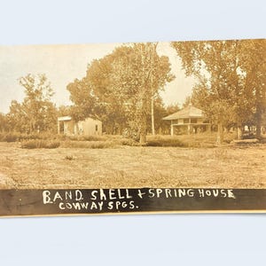 May include: Sepia-toned vintage postcard featuring two buildings in a grassy field, surrounded by trees. The text "BAND, SHELL + SPRING HOUSE, CONWAY SPGS." is printed at the bottom. The buildings appear to be simple structures, possibly a bandstand and a spring house.