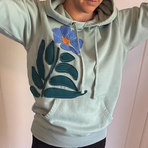 以下が含まれることがあります： 大きな花のアップリケが付いた水色のパーカー。アップリケは、オレンジ色の中心と緑の葉を持つ青い花が特徴です。パーカーには、ドローストリングフードとフロントポケットが付いています。