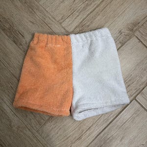 Pode incluir: Calções de tecido felpudo laranja e branco. Os calções são divididos verticalmente, com um lado laranja e o outro branco. O tecido parece macio e absorvente, sugerindo um estilo casual ou de praia.