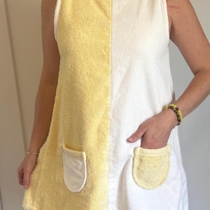 May include: A sleeveless tunic dress made of terry cloth fabric featuring a vertical color-blocked design in yellow and white. The garment includes two rounded patch pockets, with a white pocket on the yellow side and a yellow pocket on the white side.