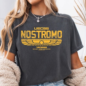 Könnte beinhalten: Dunkelgraues T-Shirt mit dem gelben Text "USCSS NOSTROMO" und einem stilisierten Emblem. Das Shirt hat einen Vintage-Look mit leicht verblichenem Aussehen. Das Design erinnert an ein Science-Fiction-Thema.