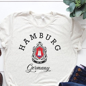 May include: A cream-coloured short-sleeve t-shirt featuring the text HAMBURG in a black arched font above a red, white, and blue crest with a castle icon, and the word Germany in black script below the crest, with the text EST. 808 inside the crest.