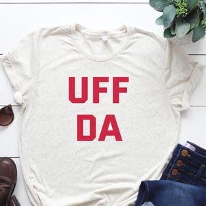 May include: A light beige t-shirt with the red text "UFF DA" is displayed on a white wooden surface. The shirt is paired with blue jeans, brown boots, and sunglasses. A small green plant is in the upper right corner.