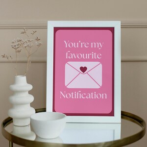 May include: A framed print with the text "You're my favourite Notification" in white on a pink background. The image includes a white envelope with a heart. A white vase and bowl sit on a gold table.