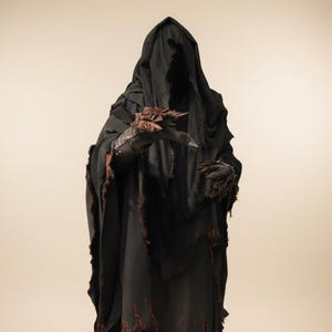 Può includere: Immagine a figura intera di una figura oscura in una veste nera con cappuccio. La veste ha bordi strappati con dettagli marroni. Le mani della figura sono adornate con accessori a forma di artiglio, il volto è nascosto dal cappuccio.