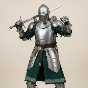 May include: A full suit of medieval armor, including a helmet, chest plate, and leg guards, is displayed. The armor is silver-colored and worn over a green tunic. A long sword is held over the shoulder. The armor is detailed and appears to be functional.