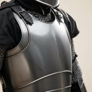 May include: Close-up of a polished steel breastplate with leather straps and buckles. The armor is part of a medieval knight's outfit, with chainmail visible on the side. The chest piece has curved edges and a reflective surface.