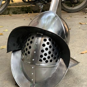 May include: A polished, silver-colored gladiator helmet. The helmet features a rounded top, a face guard with numerous small holes, and a wide, curved brim. A vertical crest rises from the top. The helmet is designed for protection and historical reenactment.