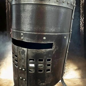 Op de afbeelding: Een verweerde, donkere metalen helm met een cilindrische vorm. De helm heeft een horizontale oogsleuf en een lager gedeelte met rechthoekige ventilatiegaten. Klinknagels bevestigen de metalen platen.