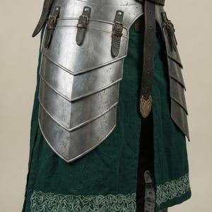 May include: Close-up of medieval armor, featuring layered metal plates and leather straps. The armor is silver with a dark leather belt and a green fabric skirt with white embroidered trim. The design suggests historical or fantasy themes.
