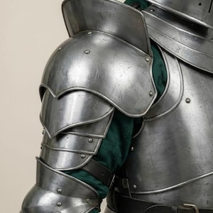 May include: Close-up of a section of medieval armor. The silver-colored metal armor includes a shoulder guard, arm protection, and chest plate. A green fabric is visible beneath the armor. The armor is secured with rivets and leather straps.