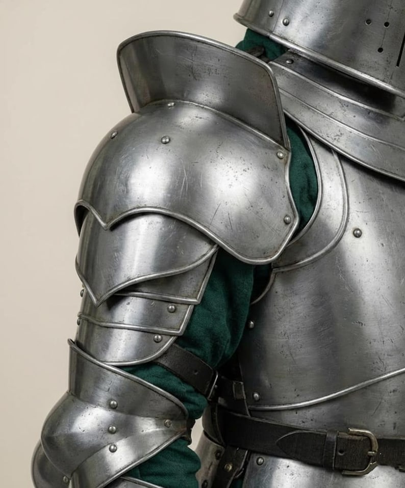 May include: Close-up of a section of medieval armor. The silver-colored metal armor includes a shoulder guard, arm protection, and chest plate. A green fabric is visible beneath the armor. The armor is secured with rivets and leather straps.