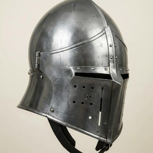 May include: A steel medieval helmet with a rounded top and a visor featuring multiple small holes. The helmet has a hinged visor and a chin strap. The metal surface shows signs of wear and tear, suggesting age and use.
