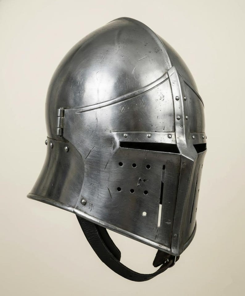 May include: A steel medieval helmet with a rounded top and a visor featuring multiple small holes. The helmet has a hinged visor and a chin strap. The metal surface shows signs of wear and tear, suggesting age and use.