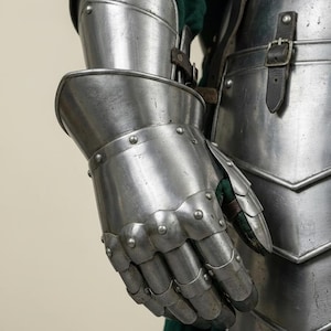 May include: Close-up of a medieval steel gauntlet, part of a suit of armor. The gauntlet is articulated, with individual plates for the fingers and wrist. Rivets secure the plates. A green fabric is visible beneath the armor.