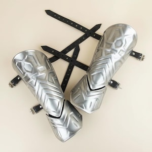 May include: A pair of silver-colored arm guards with geometric designs and black leather straps. The arm guards are made of metal and have a polished finish. The straps have buckles for secure fastening.