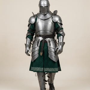 May include: Full suit of medieval armor, including a helmet with a visor, shoulder and chest plates, arm and leg protection, and gauntlets. The armor is silver-colored metal, worn over a green tunic with a decorative border. A black belt is around the waist.