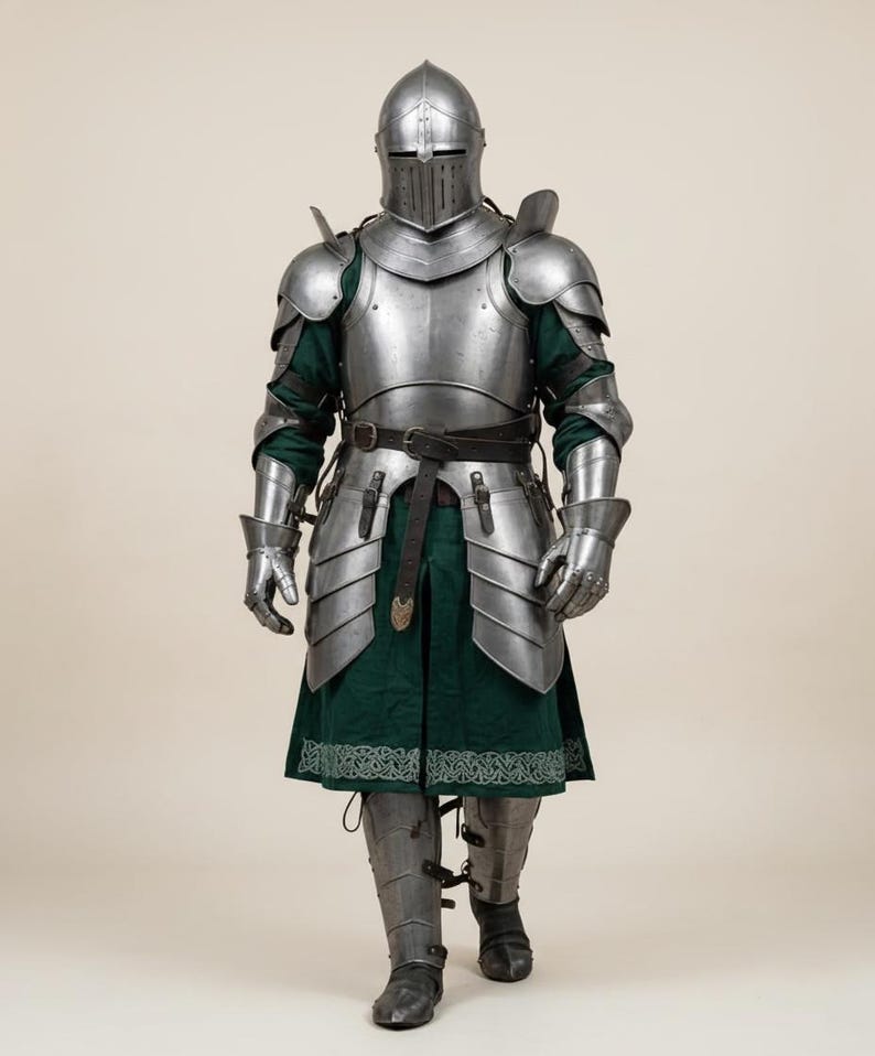 May include: Full suit of medieval armor, including a helmet with a visor, shoulder and chest plates, arm and leg protection, and gauntlets. The armor is silver-colored metal, worn over a green tunic with a decorative border. A black belt is around the waist.