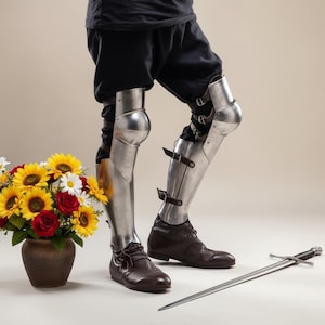 May include: A pair of polished metal leg armor with knee protection, secured by straps. The armor is worn with brown leather shoes. A bouquet of sunflowers, daisies, and red roses sits in a brown ceramic pot. A sword lies on the floor.