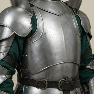 May include: Close-up of a medieval steel armor set. The armor includes a breastplate, shoulder guards, and arm protection, all riveted together. A dark leather belt with a buckle is visible. The armor is worn over a green garment.