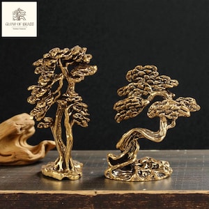 Può includere: Due sculture di bonsai in ottone. Uno è eretto, l'altro ha un tronco curvo. Gli alberi hanno un fogliame dettagliato e una corteccia strutturata. Le sculture sono su una superficie scura. Il logo "GLOW OF BRASS" è in alto a sinistra.