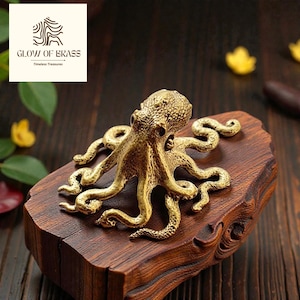 May include: A detailed brass octopus sculpture with multiple tentacles, mounted on a carved wooden base. The sculpture has a golden finish and is displayed on a dark wooden surface. The logo "GLOW OF BRASS, Timeless Treasures" is in the upper left corner.