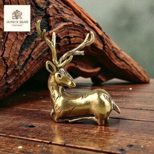 Puede incluir: Figura de ciervo de latón dorado en posición de descanso. El ciervo tiene cuernos detallados y está sobre una superficie de madera. El texto "GLOW OF BRASS" es visible en la esquina superior izquierda.