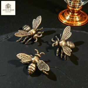 May include: Three brass bee figurines with detailed wings and body segments are arranged on a dark surface. The bees are in various poses, showcasing their intricate design. The logo "GLOW OF BRASS" is in the upper left corner.