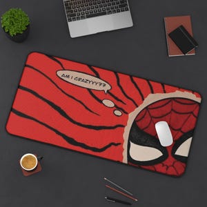 May include: A rectangular red mousepad with a comic book style Spider-Man design. The mousepad features a torn paper effect revealing Spider-Man's face and the text "AM I CRAZYYY??" in a speech bubble. A white mouse rests on the pad.