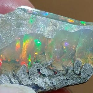 Può includere: Un'opale grezza con una vibrante gamma di colori. L'opale presenta una miscela di tonalità verdi, gialle, arancioni e blu, con un esterno ruvido e strutturato. La pietra è tenuta in mano, mostrando la sua bellezza naturale.