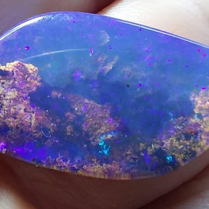 May include: A polished opal gemstone displaying a spectrum of colours. The stone has a deep blue base with flashes of purple, gold, and green. The opal's surface is smooth and reflective, highlighting its unique internal patterns.