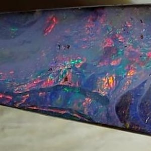 Könnte beinhalten: Ein polierter, länglicher Opal zeigt ein faszinierendes Farbspiel. Die Oberfläche des Steins ist ein tiefes, durchscheinendes Blau mit lebendigen Blitzen in Rot, Grün und Gold. Das einzigartige Muster und die Farbvariationen des Opals machen ihn zu einem auffälligen Edelstein.