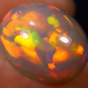 May include: A close-up of a polished, oval-shaped opal gemstone. The opal displays a vibrant play-of-color, with flashes of orange, yellow, green, and hints of blue. The gemstone has a translucent appearance, with a smooth, reflective surface.