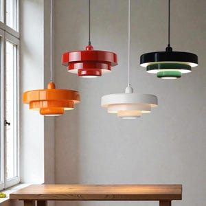 May include: Four contemporary pendant lights in red, orange, white, and black. Each light has a tiered design with a white interior, suspended from a thin cord. The lights hang above a wooden table, creating a stylish interior.