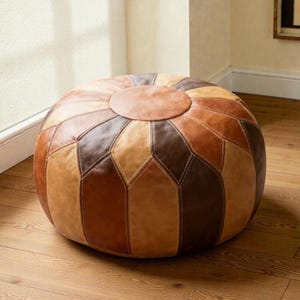 Puede incluir: Un puf redondo de cuero con diseño de patchwork en tonos marrones y beige. La parte superior es un círculo marrón claro liso. El puf es una opción de asiento bajo, a nivel del suelo, con un diseño geométrico y costuras visibles.