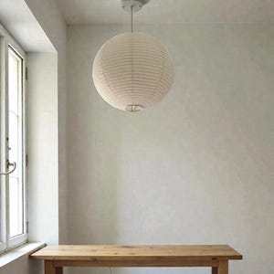 May include: A spherical, cream-coloured paper lantern hangs from the ceiling. The lantern is suspended above a wooden table. The room has a minimalist aesthetic with a neutral colour palette and natural light from a nearby window.