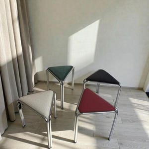 May include: Four modern stools with triangular seats in various colors: light gray, dark green, black, and red. Each stool has a silver metal frame. The stools are arranged on a light wood floor.